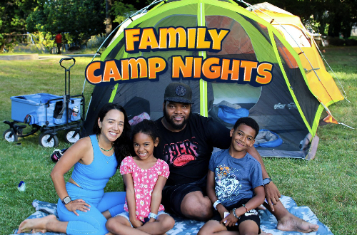 Family of four sitting on a blue and white picnic blanket in-front of their green and grey tent