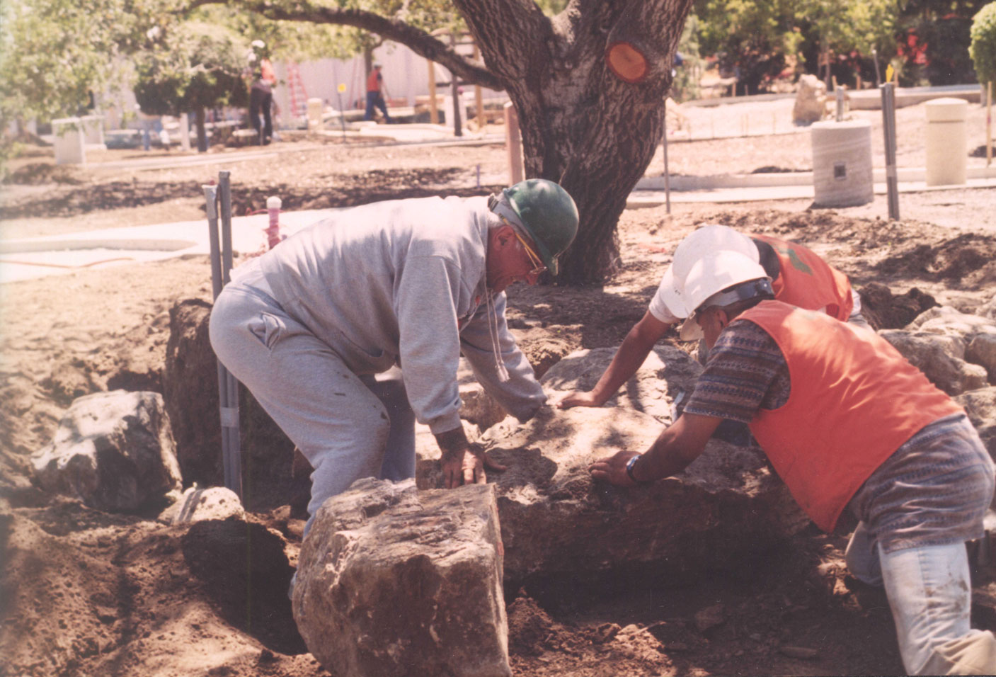 Three men in grey and orange clothes moving a rock near a tree