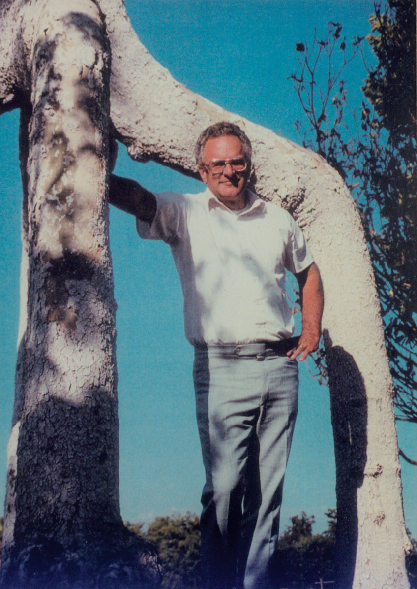Man in a white shirt and blue jeans standing between the Four-Legged Giant circus tree