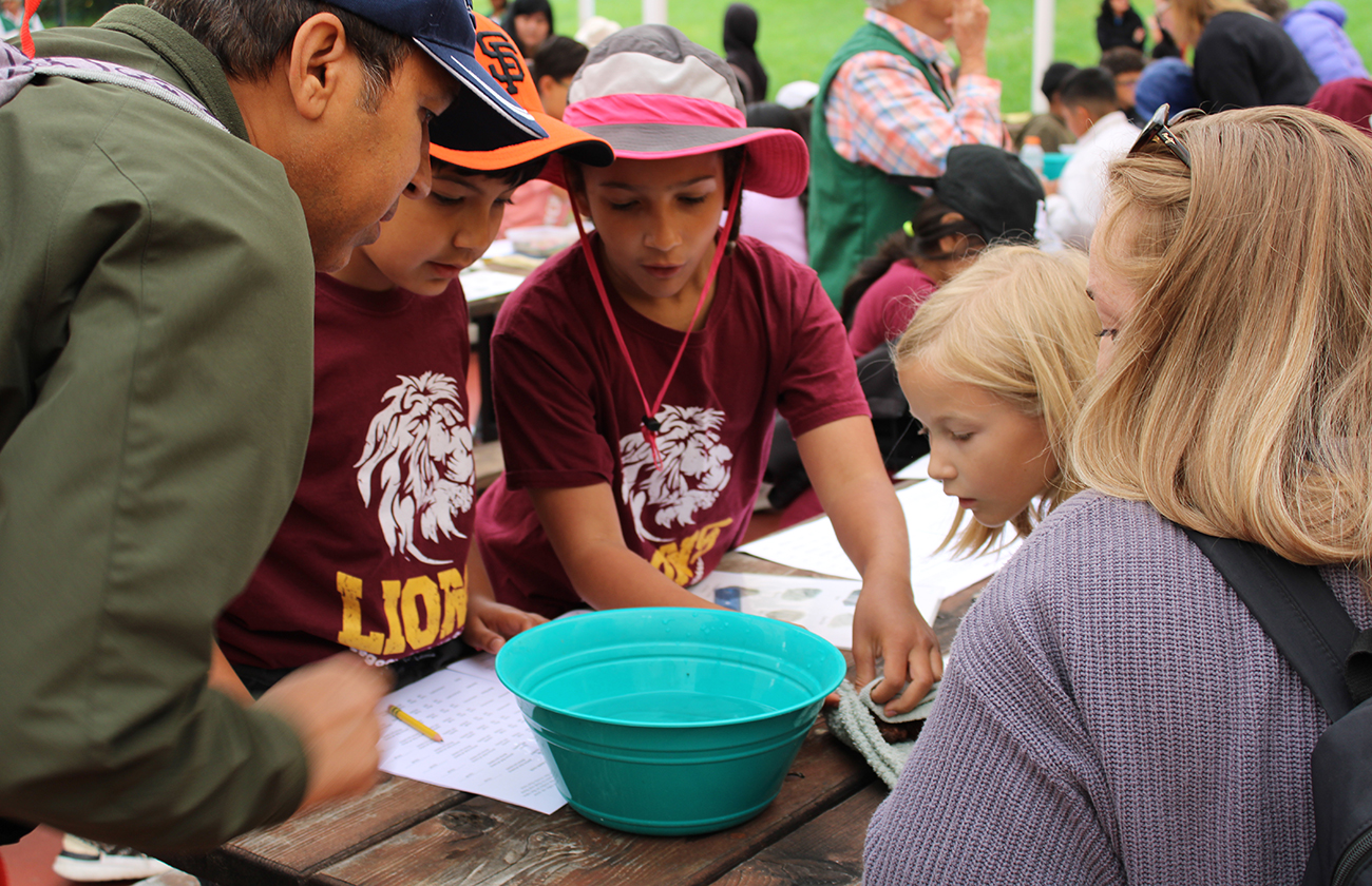 Group of children wearing maroon shirts looking in amazement into a turquoise bowl with water