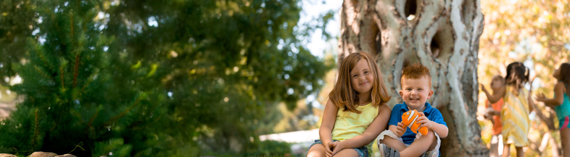 A young girl in a yellow sleeveless shirt and young boy in a blue collar shirt holding a goldfish stuffed animal smiling while sitting in front of a Basket Tree