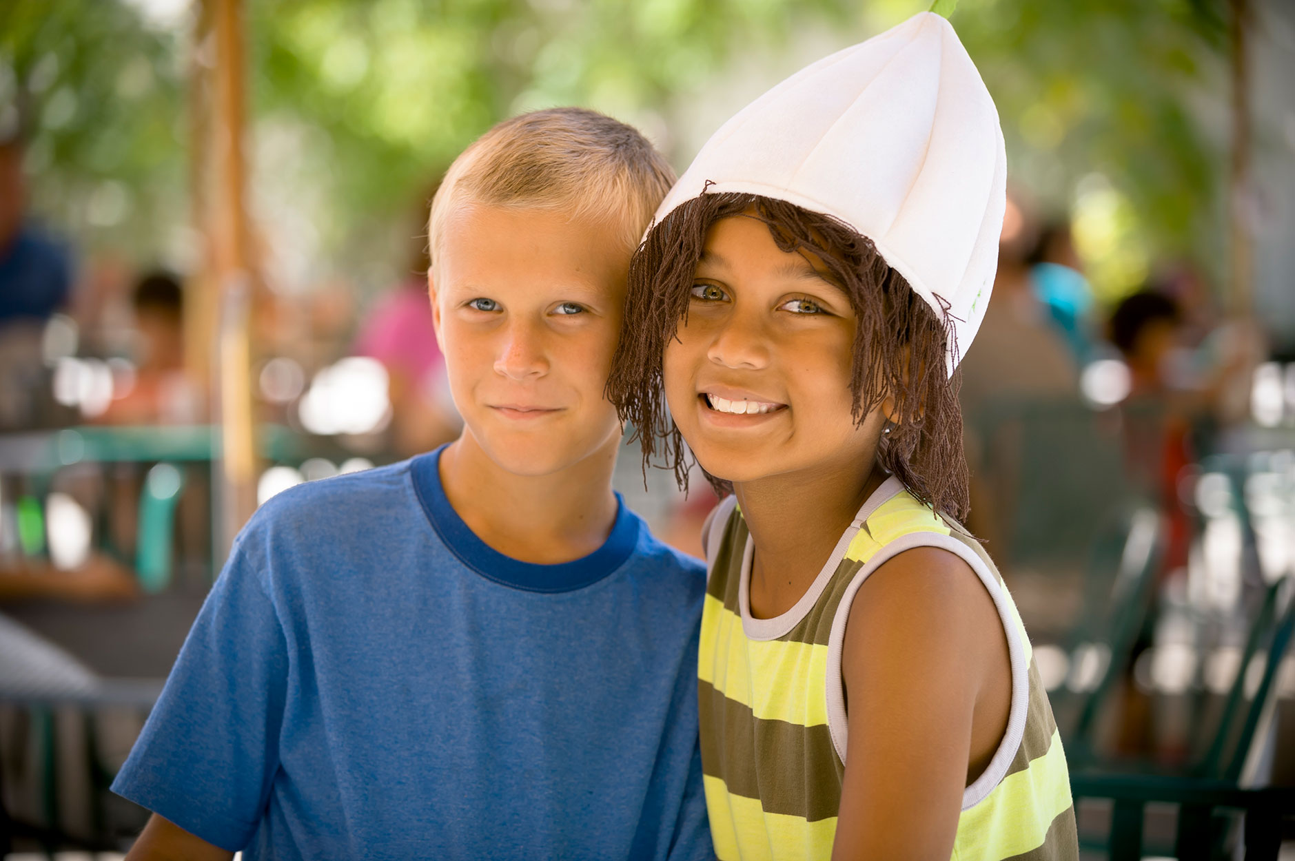 Boy in a blue t-shirt standing and smiling next to his friend in a striped yellow and brown tank top wearing a garlic hat
