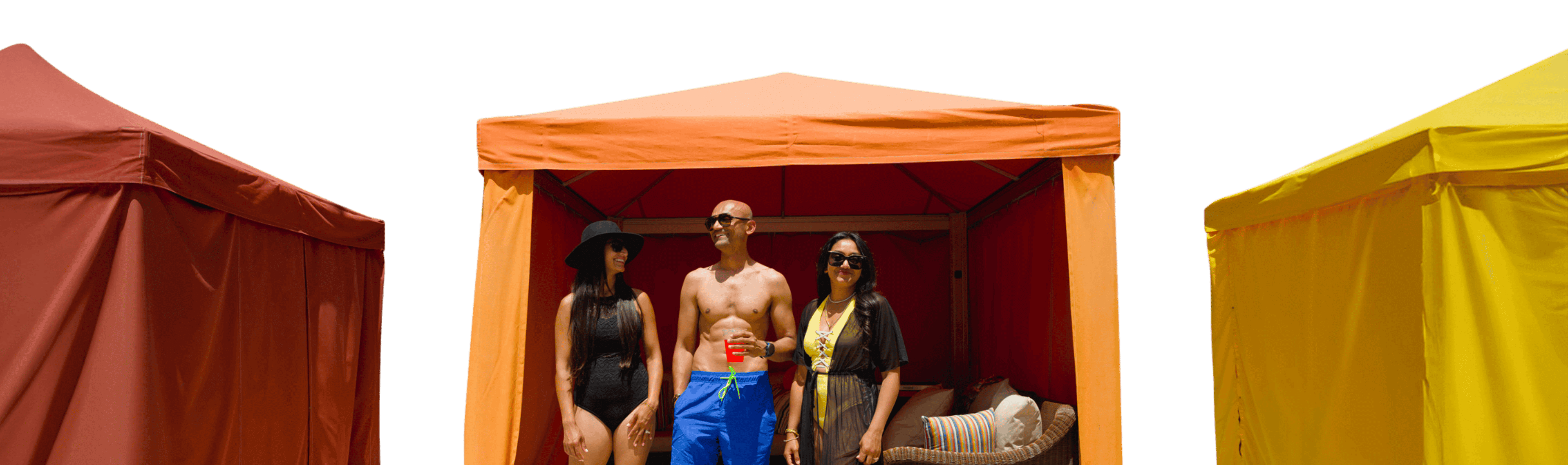 Man in blue swim trunks standing with a woman in a black swimsuit and another woman in a yellow swimsuit with a black sheer cover in front of an orange cabana