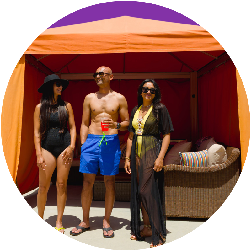 Man in blue swim trunks standing with a woman in a black swimsuit and another woman in a yellow swimsuit with a black sheer cover in front of an orange cabana