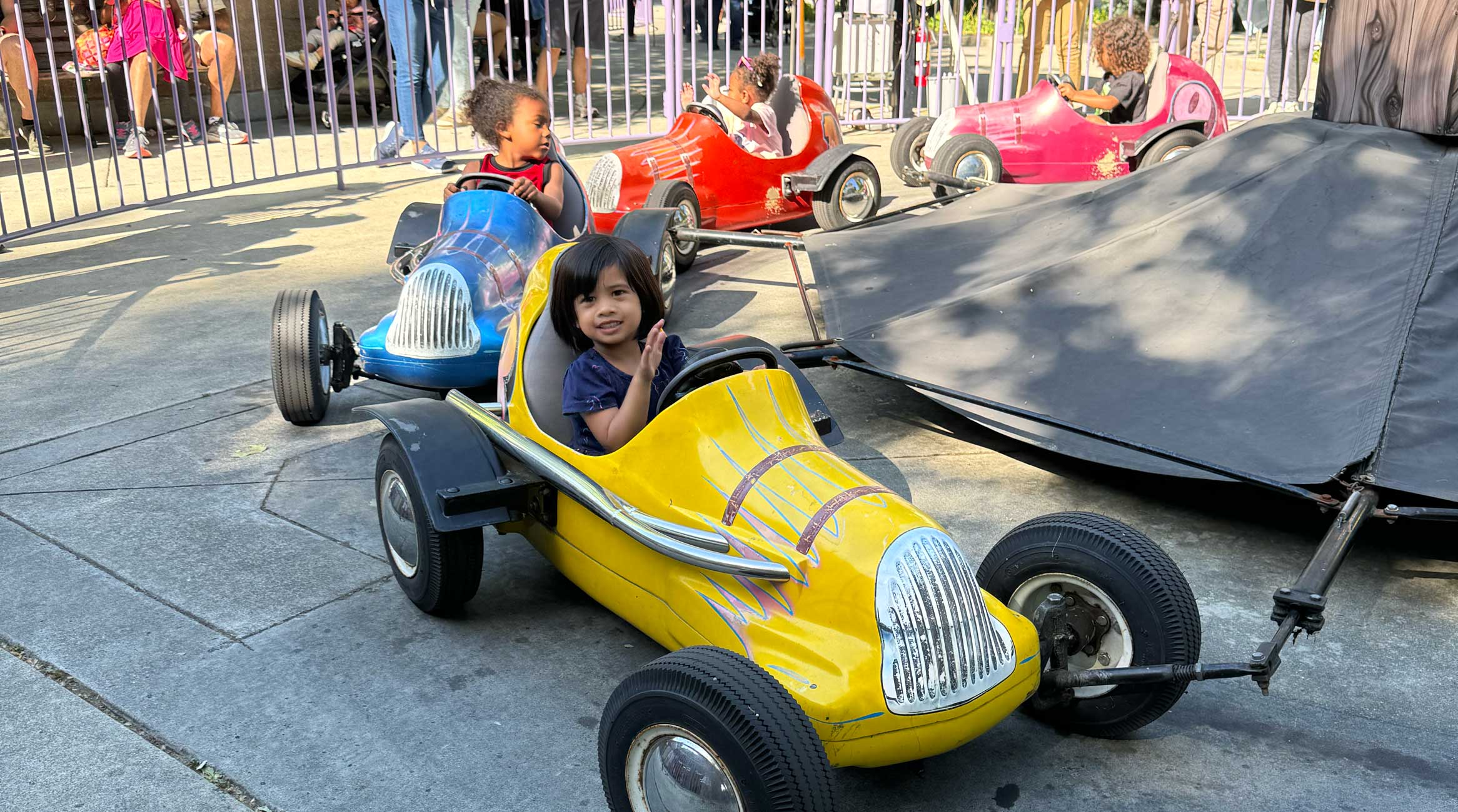 Little girl in a blue shirt waving while in her yellow car on the Pit Stop Racers ride