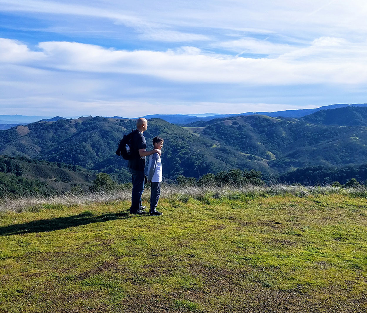 Father placing his hand on his son's shoulder while hiking on a green hill overlooking the city