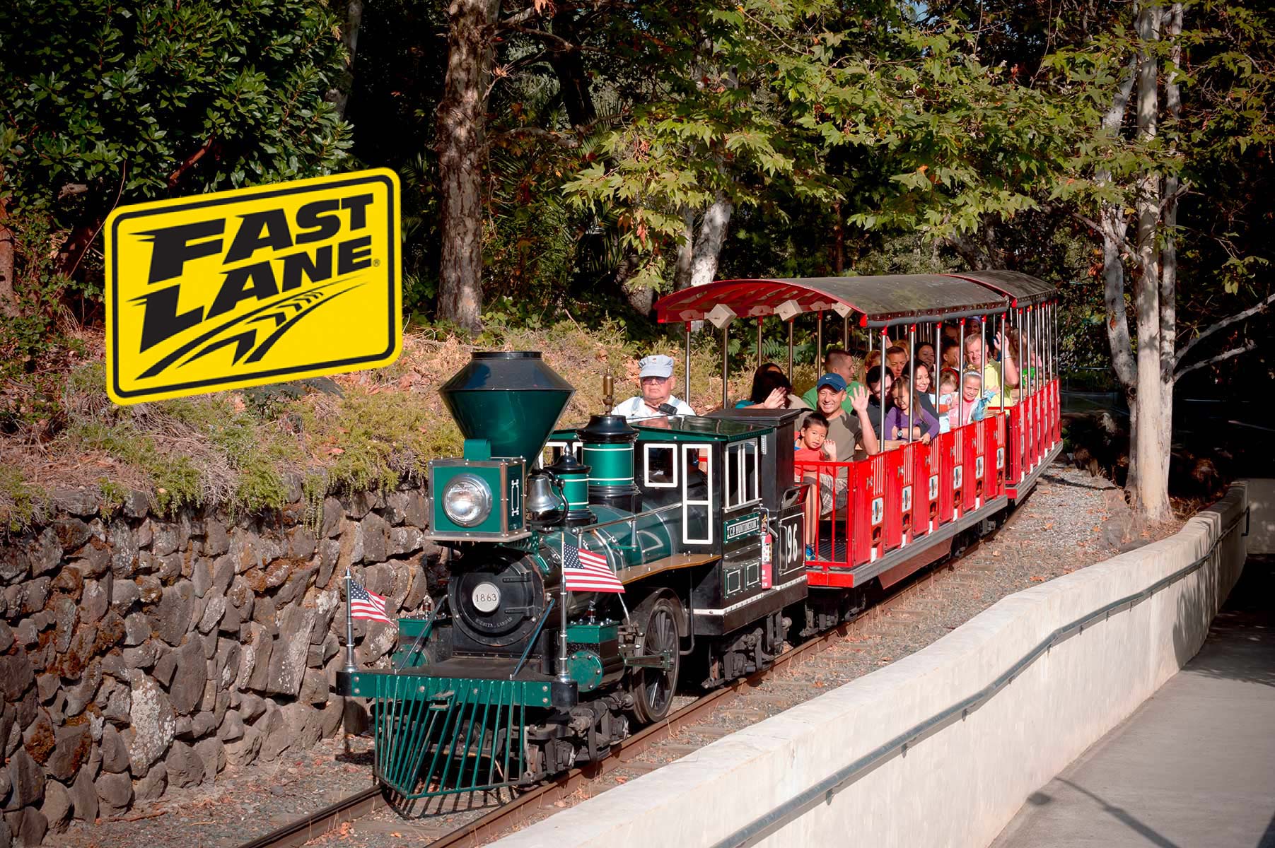 Green train with two American flags going down the tracks with large green trees around it
