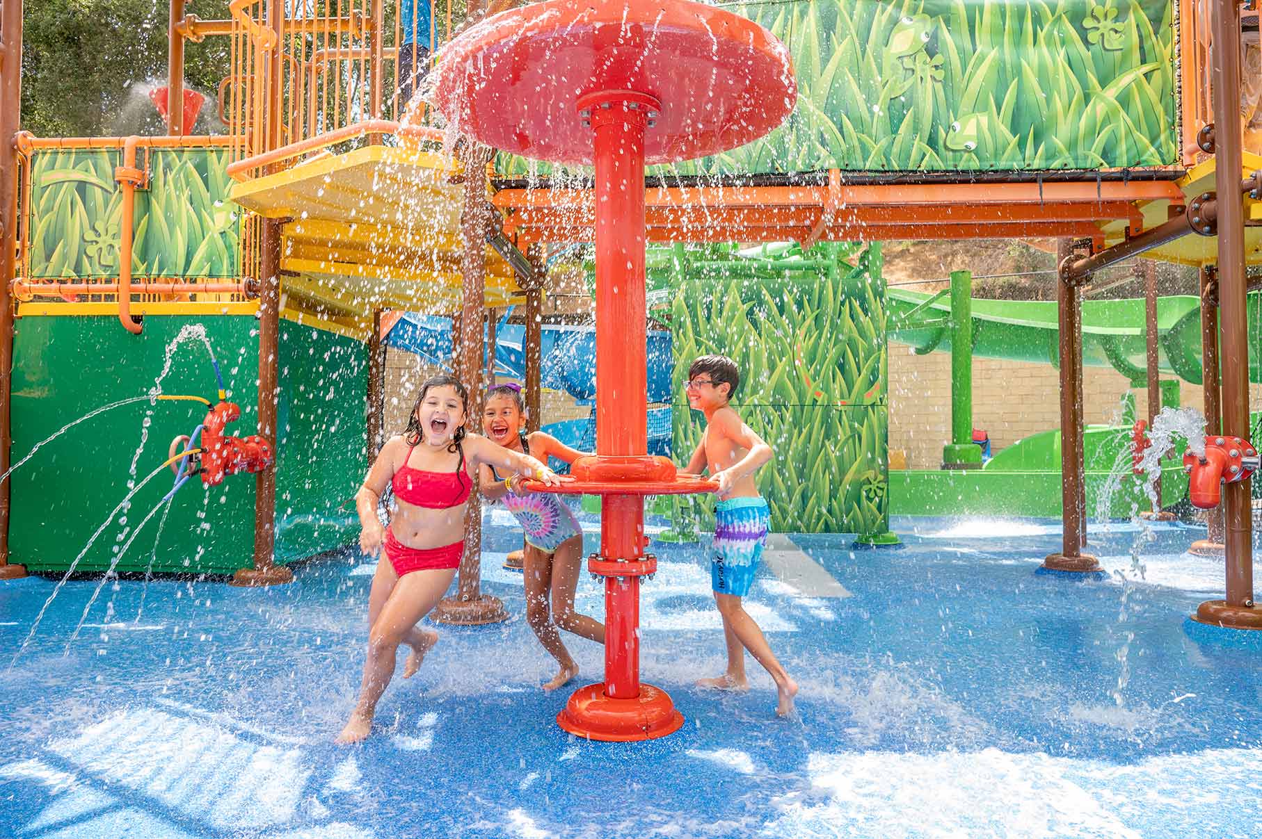 A girl in a red two piece swimsuit, girl in a one piece tie dye swimsuit and a boy in tie dye trunks play at Water Oasis