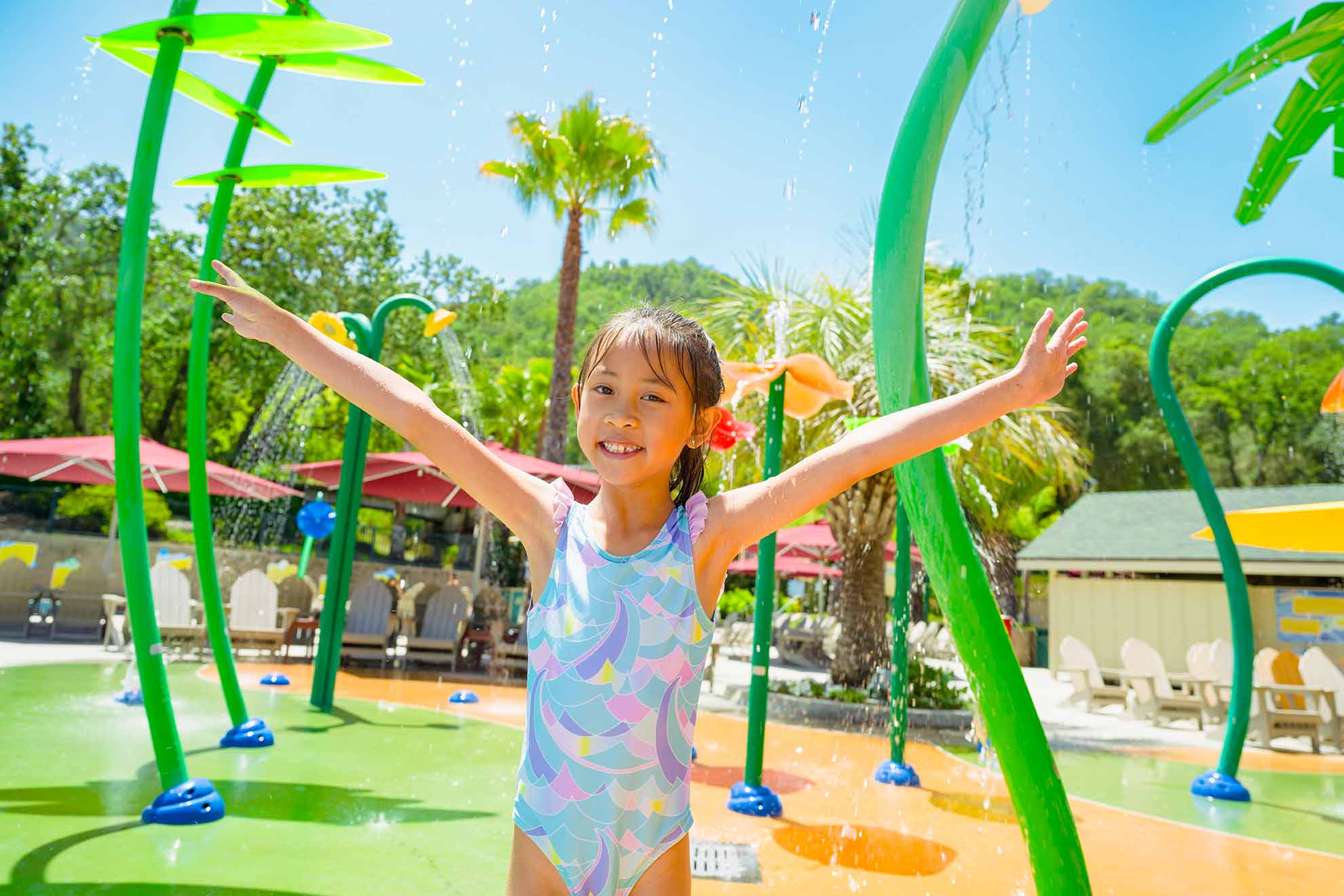 Young girl in a blue, pink, and yellow bathing suite with her arms up and smiling at Water Oasis