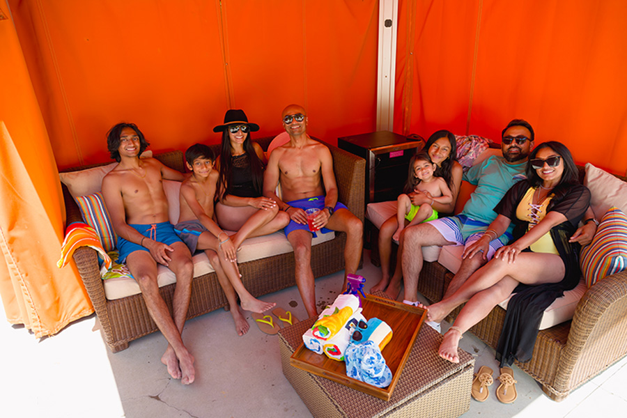 Large family in swimsuits relaxing on beige outdoor seating inside an orange cabana at Water Oasis