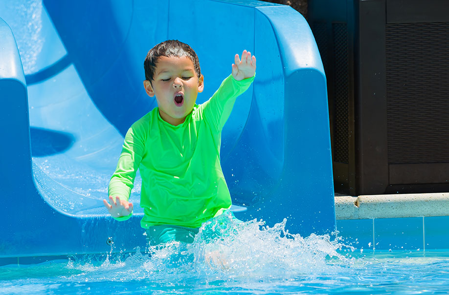 Young boys in bright green shirt with his hands in the air going down a blue slide at water oasis.