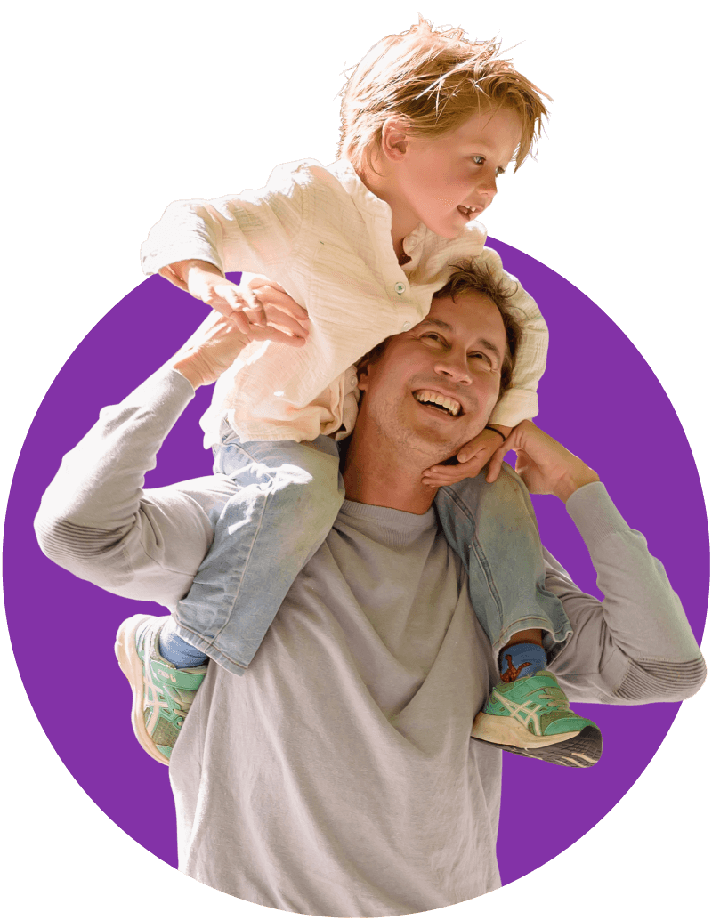 Dad smiling while wearing a light grey shirt carrying his son that is wearing a cream colored shirt.