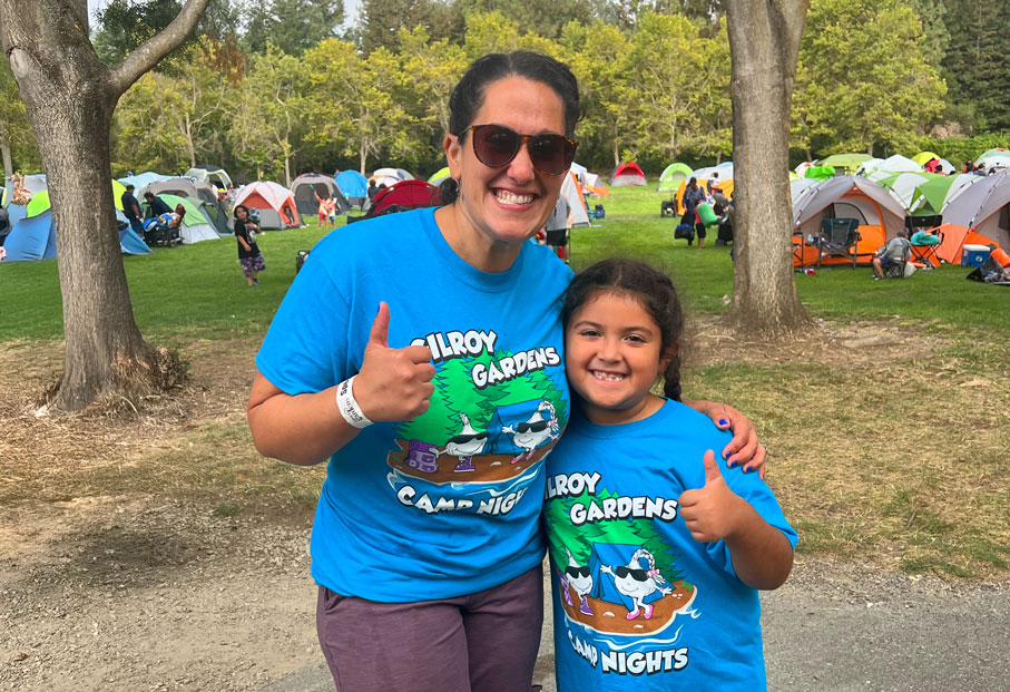 Mom and daughter in matching blue Gilroy Gardens Camp Nights shirts smiling with their thumbs up