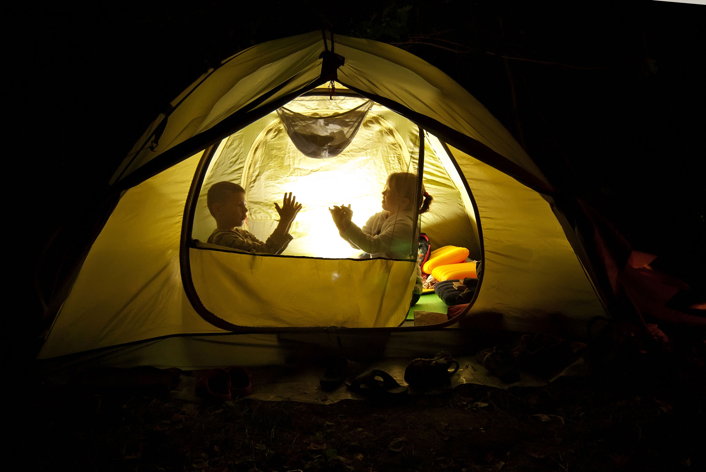 A young boy and girl inside of a yellow tent at night with a light on making animal shapes with their hands