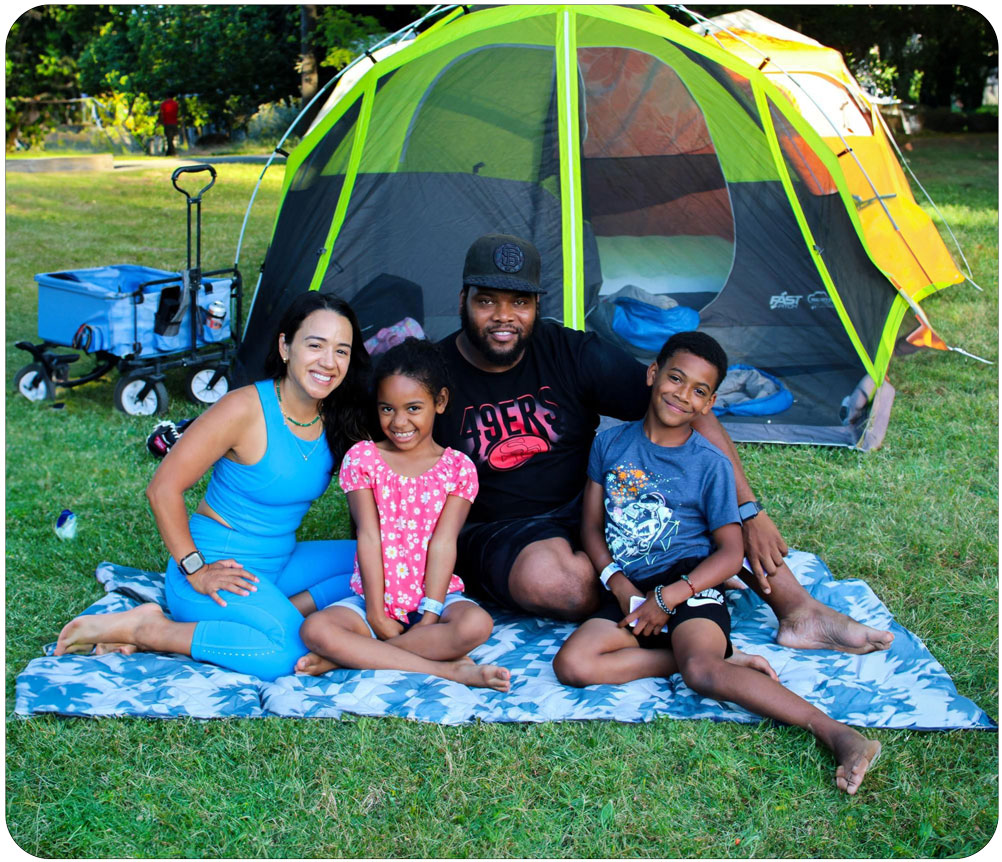 Family of four sitting on a blue and white picnic blanket in-front of their green and grey tent