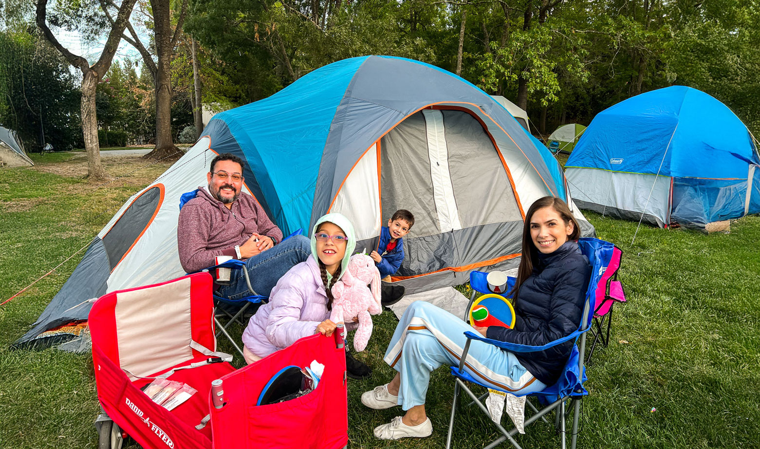 Family of four sitting on lawn chairs in-front of their blue, orange, and grey tent