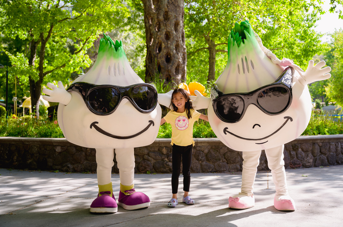 Young girl in a yellow shirt and black pants in the middle of Gil and Roy mascots having fun in front of tree