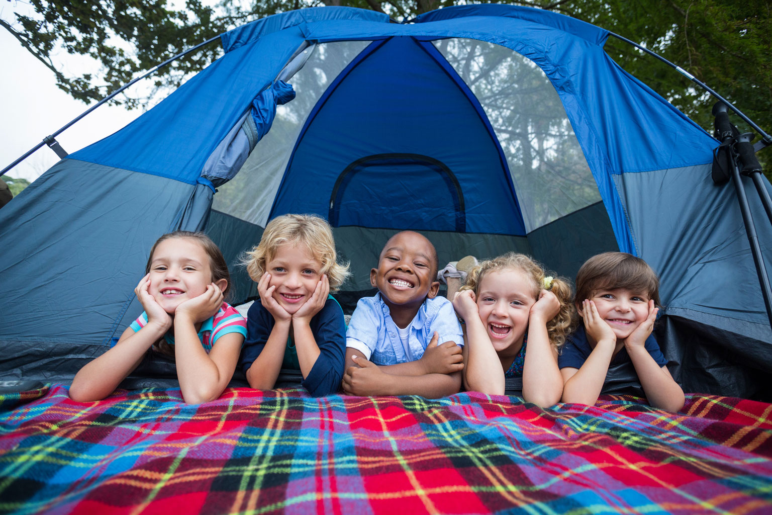 Young boys and girls smiling while laying on a red and yellow blanket in front of a blue tent