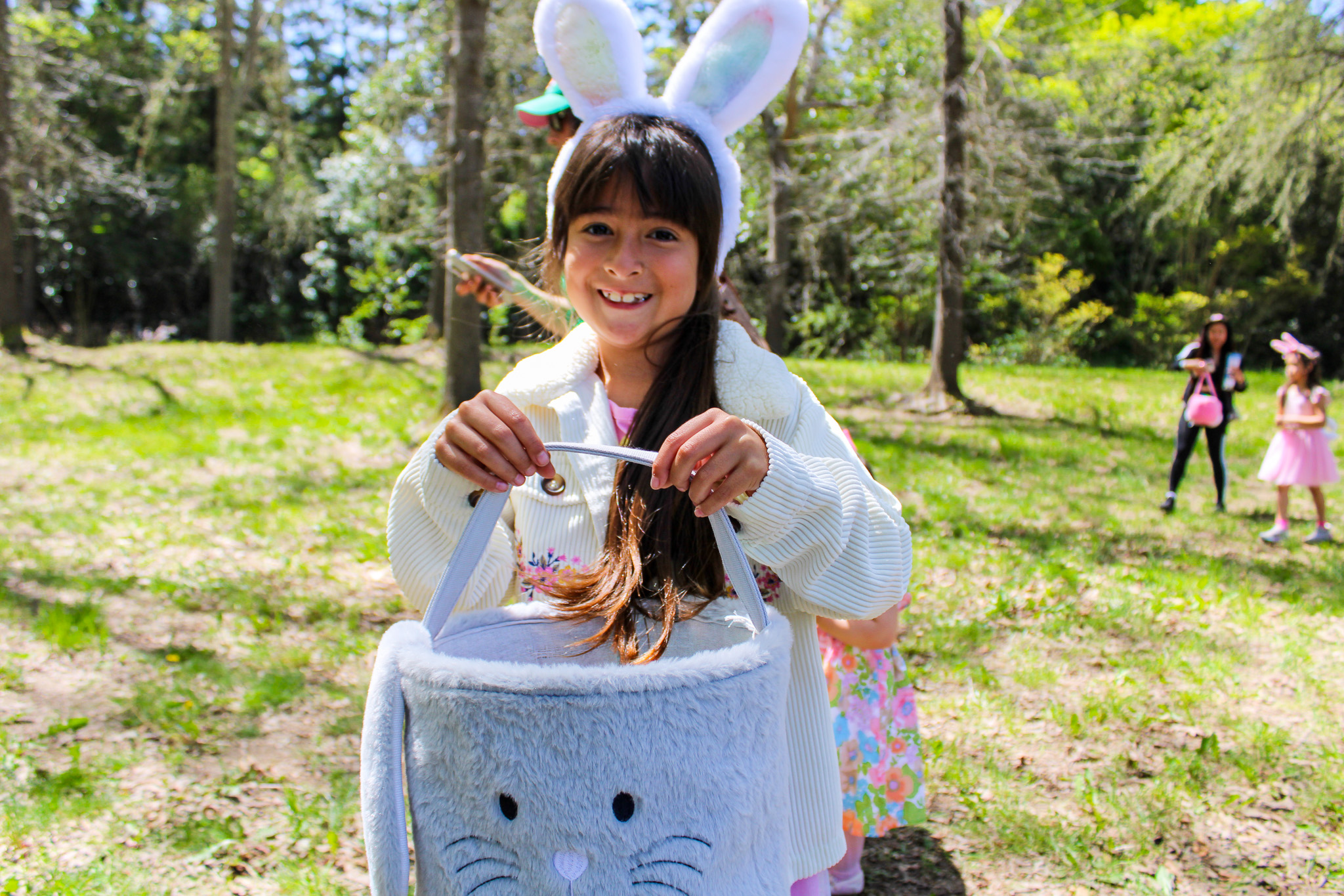 Little girl smiling and wearing a rabbit ear headband while holding a gray rabbit-themed basket.
