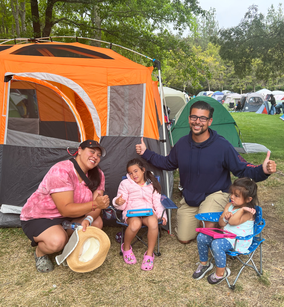 Family of four sitting on lawn chairs in-front of their orange and grey tent