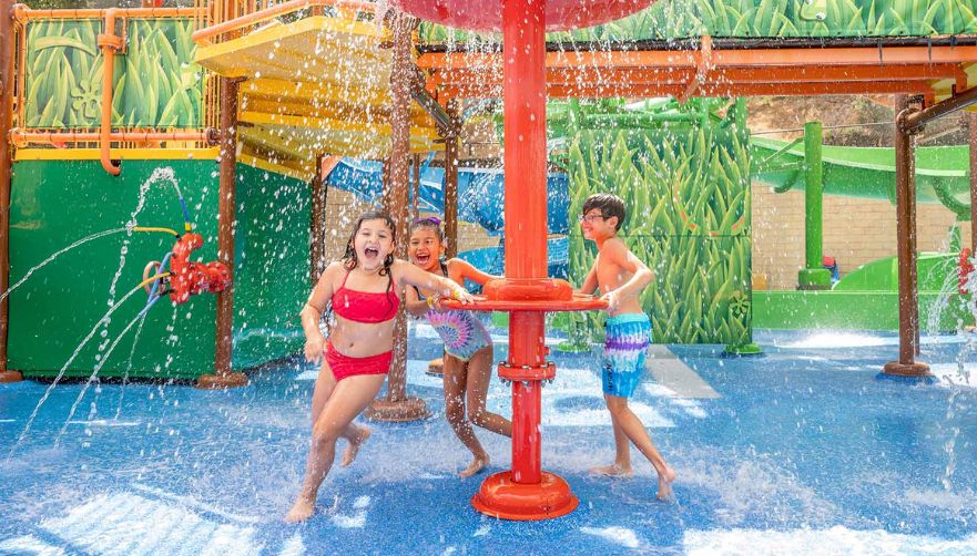 Little girl in a two piece red swimsuit, little girl in a one piece tie-dye swimsuit, and little boy in tie-dye swim trunks smiling and laughing while they get wet