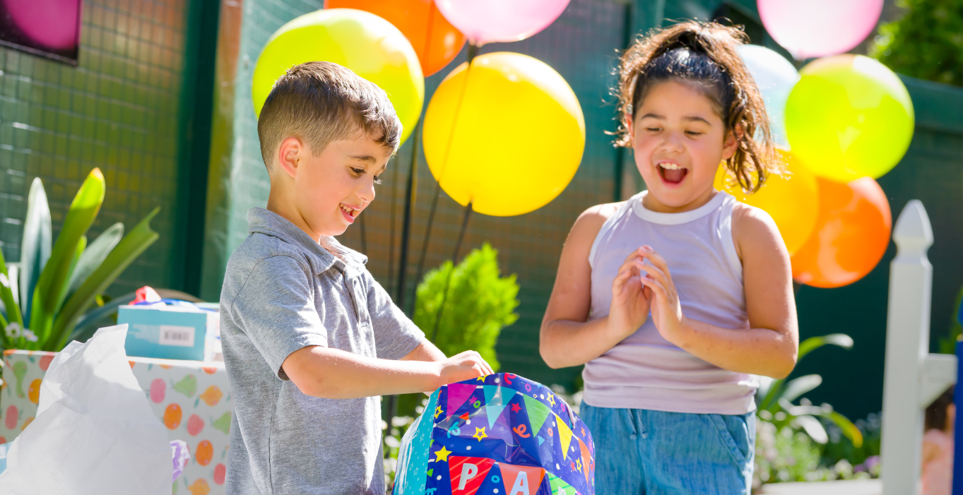 A young boy smiling while opening a birthday present and a young girl excited to see the gift