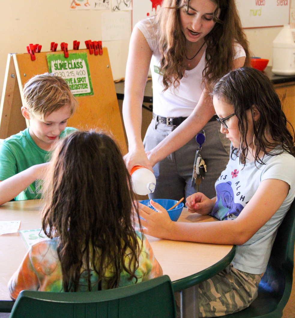 Green Barn associate assisting three children by pouring glue into a blue bowl.